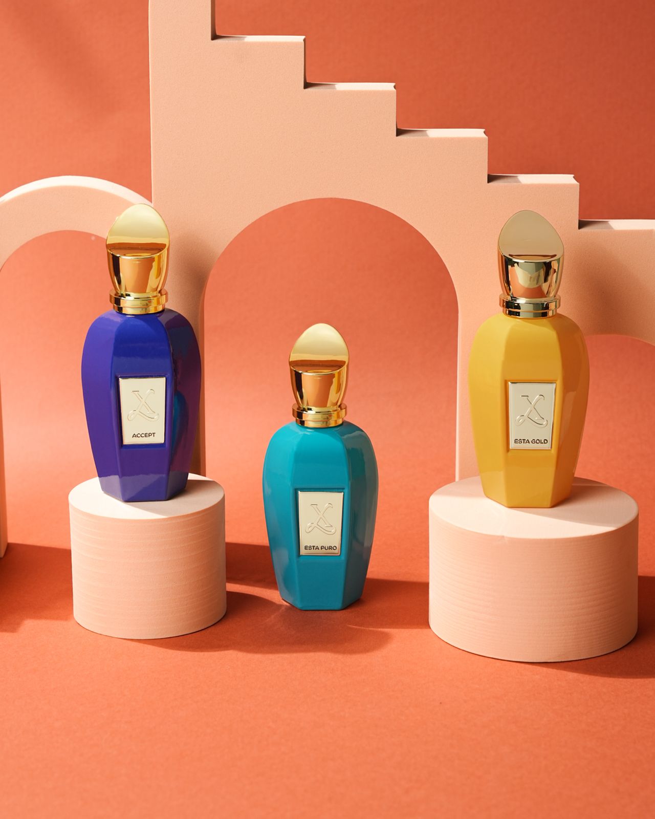 Three colorful perfume bottles on cylindrical stands against a geometric pink and orange background.